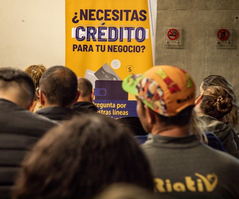 Se observa un grupo de persoans sentadas de espalda y al fondo un carytel que dice "¿Necesitas crédito para tu negocio?"